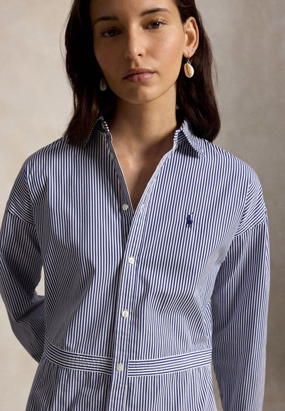 Γυναίκα που φοράει πουκάμισο με κάθετες μπλε και λευκές ρίγες και μικρό κεντημένο λογότυπο, με σκουλαρίκια σε σχήμα κοχυλιού, στέκεται μπροστά σε απλό φόντο.