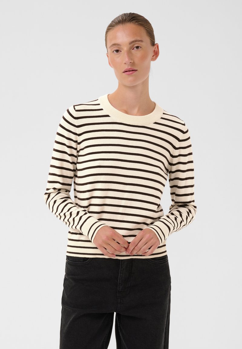 Femme aux cheveux tirés en arrière, portant un pull blanc à manches longues avec des rayures horizontales noires et un pantalon noir, debout devant un fond uni.