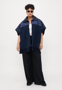 Personne portant des lunettes de soleil, un poncho zippé frangé bleu marine avec des épaules matelassées, un haut blanc et un pantalon noir ample, se tenant contre un fond uni.
