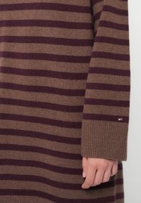 Πλεκτό πουλόβερ σε καφέ και μπορντό ρίγες. Διαθέτει ριμπ μανσέτα και μικρή λεπτομέρεια λογότυπου κοντά στον καρπό. Μαλακή υφή, μακριά μανίκια.