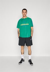 Zöld sportpóló sárga "ADIDAS" felirattal, fekete sorttal párosítva. Ezenkívül fekete sportcipő és fehér sportzokni is található.