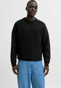 Svart stickad tröja med ribbat rundhals och långa ärmar, kombinerad med blå högmidjade jeans. Texturerad tyg, avslappnad passform.