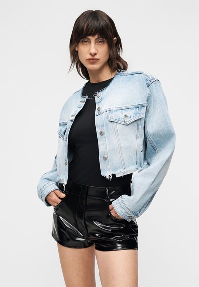 Νεαρό άτομο με σκούρα μαλλιά μέχρι τον ώμο, φορώντας ένα κοντό ανοιχτόχρωμο τζιν μπουφάν, μαύρο τοπ και γυαλιστερά μαύρα σορτς με τα χέρια στις τσέπες.