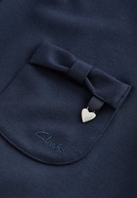 Marine stof met een zak met een strikdetail. Zilveren hartvormige bedel met de gravure "Clark" en geborduurd logo nabij de zak.