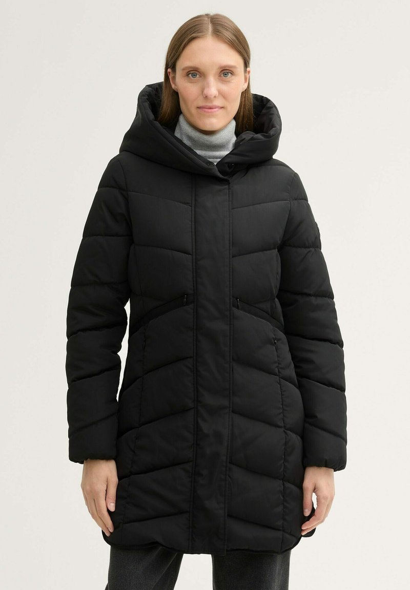 Manteau rembourré noir matelassé avec une grande capuche, un devant zippé et deux poches latérales. Présente une texture lisse et un design mi-long.