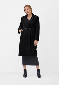 Cappotto nero doppiopetto con revers a punta, due tasche frontali e tessuto testurizzato, abbinato a una gonna grigia fino al ginocchio e stivaletti neri.