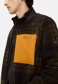 Fleecejacke mit Karomuster in Dunkelbraun und Schwarz. Verfügt über eine auffällige orangefarbene Brusttasche mit Logo und einen hohen Kragen.