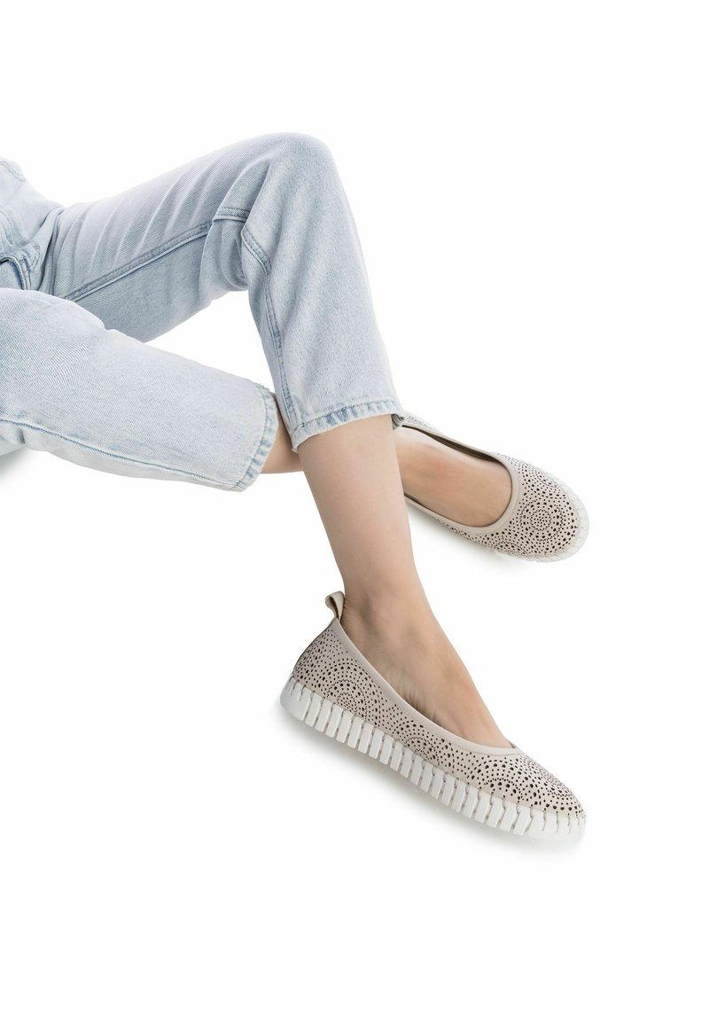 Zapatos slip-on beige perforados con una suela de goma blanca texturizada y un patrón decorativo de cortes en la parte superior. Diseño ligero y flexible.