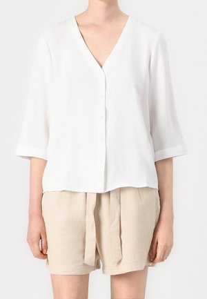 Kobieta ubrana w białą bluzkę z dekoltem w kształcie litery V, zapinaną na guziki, z rękawami do połowy przedramienia oraz beżowe szorty z materiału z wiązanym paskiem w talii.