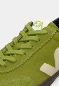 Sapatilha verde em camurça com atacadores texturizados, detalhes em creme e sola em borracha castanha. Marca em destaque na língua.