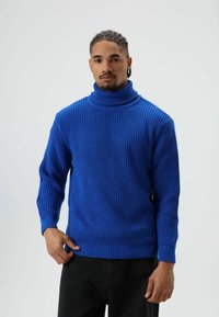 Blauwe coltrui met een geribbelde textuur, lange mouwen en een losse pasvorm, gecombineerd met zwarte broek.