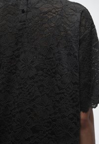 Μαύρο δαντελένιο ύφασμα με λουλουδάτο σχέδιο σε ρούχο με κοντά μανίκια, που δείχνει το μισό χέρι και τον ώμο με λαιμόκοψη με κουμπιά.