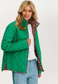 Kobieta w zielonej pikowanej kurtce z brązowo-białym wzorem w kropki, białej koszuli i jasnoniebieskich dżinsach, uśmiechająca się i patrząca w lewo.