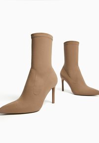 Ruskeat nilkkurit, joissa korkea piikkikorko ja sileä kangaspinta. Mallissa on vartalonmyötäinen leikkaus ja kärkiosa on terävä kärki, jossa näkyvät ompeleiden korostukset.