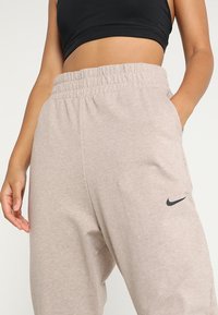 Pantalones de chándal gris claro con cintura elástica y bolsillos laterales. Presenta un logo de swoosh negro de Nike en el muslo izquierdo. Textura de tela suave.