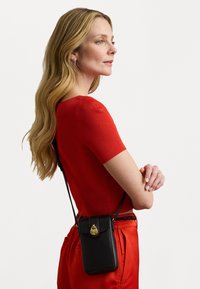 Svart axelremsväska med en guldklaff, rektangulär form, slät yta. Bärs med en röd ribbad tröja och matchande röda byxor.