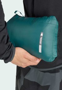 Teal puffy tas, gemaakt van lichtgewicht materiaal, heeft een handvat aan de bovenkant, een ritssluiting en een merklabel. Dicht bij een zwart kledingstuk gehouden.
