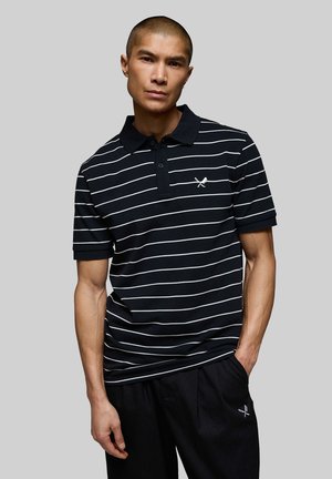 Mand iført sort og hvid stribet polo og sorte bukser med krydset bat og bold-logo på bryst og lår, stående mod grå baggrund.