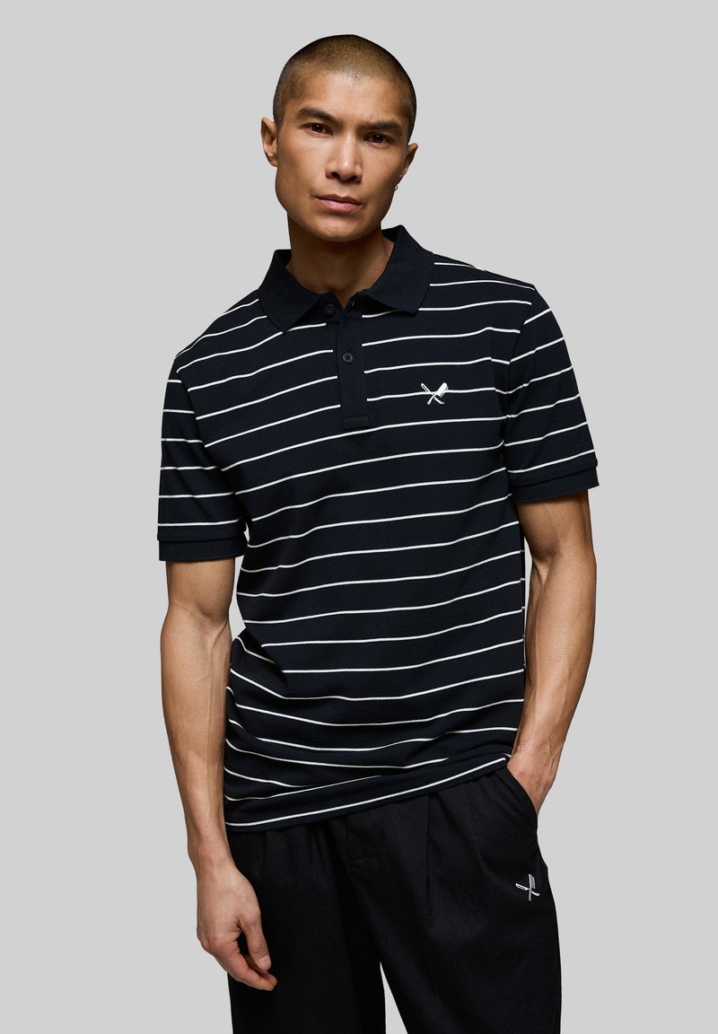 Homme portant un polo rayé noir et blanc et un pantalon noir avec un logo de batte et balle croisées sur la poitrine et la cuisse, debout devant un fond gris.