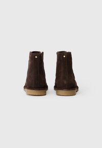 Botine din suede maro, cu un profil subțire, având o suprafață texturată, talpă netedă de culoare bej și accente metalice subtile pe spate.