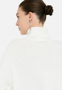 Maglione bianco a collo alto in maglia con texture a coste, caratterizzato da un alto colletto e dettagli di cucitura sottili. Orecchini a catena argentati visibili.