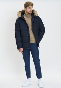 Giacca puffer blu navy con cappuccio in pelliccia sintetica, texture trapuntata, tasche frontali, abbinata a un dolcevita beige e jeans blu, completata con sneakers bianche.