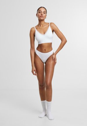 Weißes Bralette und Bikini-Set mit Spitzenbesatz. Glatter Stoff, figurbetontes Design und minimalistischer Stil. Model steht barfuß mit den Händen in den Hüften.