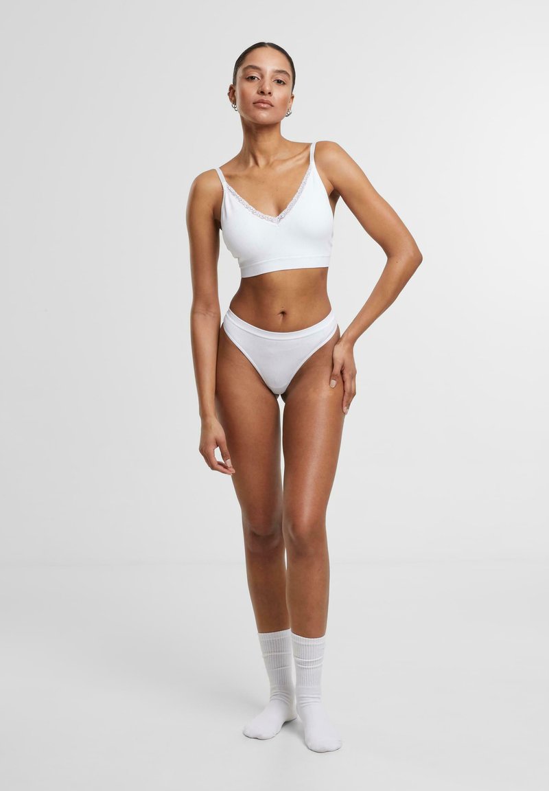 Valkoinen bralette ja bikinit-setti pitsireunuksella. Suloinen kangas, istuva muotoilu ja minimalistinen tyyli. Malli seisoo paljain jaloin, kädet lantiolla.