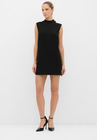 Vestido negro sin mangas con un alto escote, abertura lateral y botones en el hombro. Combinado con zapatos de tacón alto negros. Textura de tela suave.