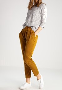 Grå långärmad tröja med strukturerat tyg och blommönster på ärmarna, kombinerad med senapsgula avsmalnande byxor och vita slip-on skor.