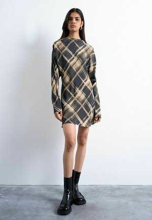 Kvinde iført en langærmet, knælængde kjole med beige og sort ternet mønster, kombineret med sorte glansfulde ankelstøvler.