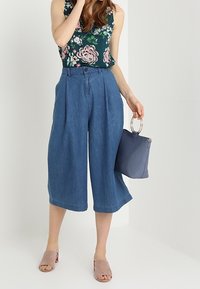 Culottes en jean à taille haute, avec des plis et une jambe large. Associés à un haut sans manches à motifs floraux et des sandales à glisser gris doux. Tenant un sac à main bleu.