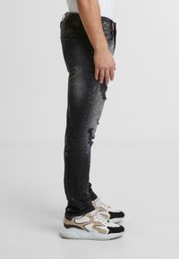 Czarne dżinsy o zwężanym kroju z przetarciami, wzorem w plamy i klasycznym pięcio kieszeniowym stylem. Noszone z białymi i czarnymi trampkami.