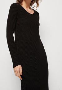 Robe noire côtelée à manches longues avec un décolleté rond, présentant des rayures verticales et une silhouette ajustée. Texture douce et longueur moyenne.