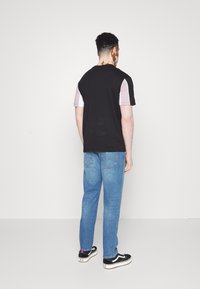 Camisa preta e rosa de mangas curtas com um corte descontraído, combinada com jeans azuis e ténis pretos com detalhes brancos, vista pela parte de trás.