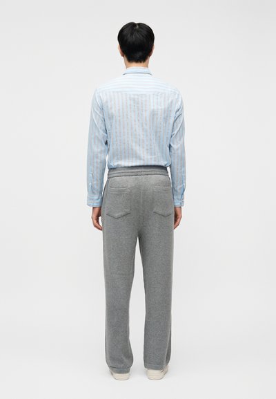 Άνδρας στέκεται γυρισμένος προς τα πίσω φορώντας γαλάζιο πουκάμισο με ρίγες, γκρι παντελόνι με φαρδιά μπατζάκια και πίσω τσέπες, καθώς και λευκά αθλητικά παπούτσια, σε απλό φόντο.