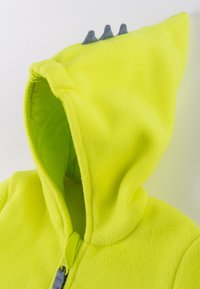 Yellow fleece hoodie with a lined hood, featuring three gray fabric spikes on top and a zippered front closure. Soft texture.