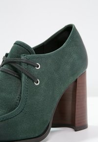 Chaussure Oxford en cuir vert avec une finition texturée, dotée de lacets gris, d'œillets argentés et d'un talon en bois robuste.