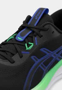 Chaussure de sport noire avec une tige texturée, des accents bleus et verts, et une semelle verte contrastante. Lacets intégrés aux œillets pour un meilleur maintien.