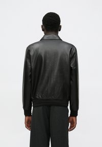 Casaco bomber preto em pele com textura suave, punhos e gola canelados. Design simples e minimalista, sem marcas ou padrões visíveis.