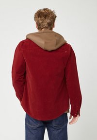 Uomo con capelli castano chiaro che indossa una giacca di velluto a coste rossa sopra un hoodie marrone e jeans blu, in piedi con la schiena rivolta in avanti.