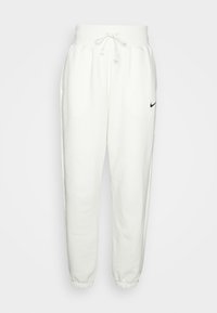 Bílé Nike tepláky s elastickými manžetami, stahovací pas, boční kapsy a malé černé logo swoosh na levém stehně.