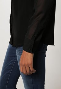 Black sheer shirt featuring long sleeves with buttoned cuffs, paired with blue denim jeans, highlighting a smooth fabric texture.