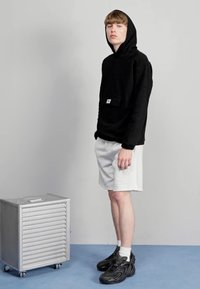 Felpa nera con cappuccio e tasca a marsupio, abbinata a pantaloni corti grigi. Scarpe da ginnastica nere e una scatola di metallo accanto. Sfondo chiaro.