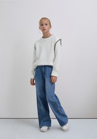 Joven de pie, vistiendo un suéter blanco con ribete negro, jeans anchos azules con patrones de estrellas y zapatillas blancas sobre un fondo liso.