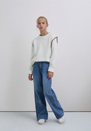 Joven de pie, vistiendo un suéter blanco con ribete negro, jeans anchos azules con patrones de estrellas y zapatillas blancas sobre un fondo liso.