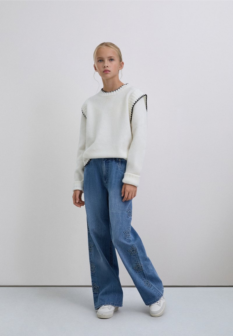 Joven de pie, vistiendo un suéter blanco con ribete negro, jeans anchos azules con patrones de estrellas y zapatillas blancas sobre un fondo liso.