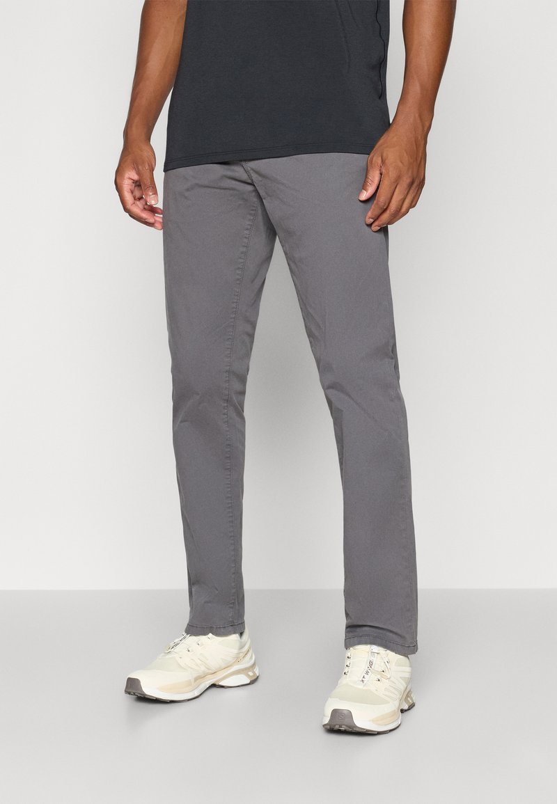 Mann in grauen Slim-Fit-Hosen, schwarzem T-Shirt und cremefarbenen Sportschuhen vor einem schlichten Hintergrund.