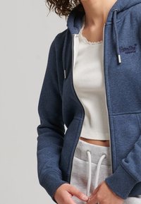 Sudadera con cremallera azul marino de tela suave, con un bolsillo canguro y un logo en el pecho, combinada con una camiseta blanca de canalé y pantalones de chándal grises.