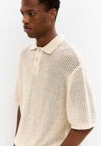 Beige, stickad polo skjorta med korta �ärmar, med en texturerad meshdesign och tre knappar vid knäppningen.